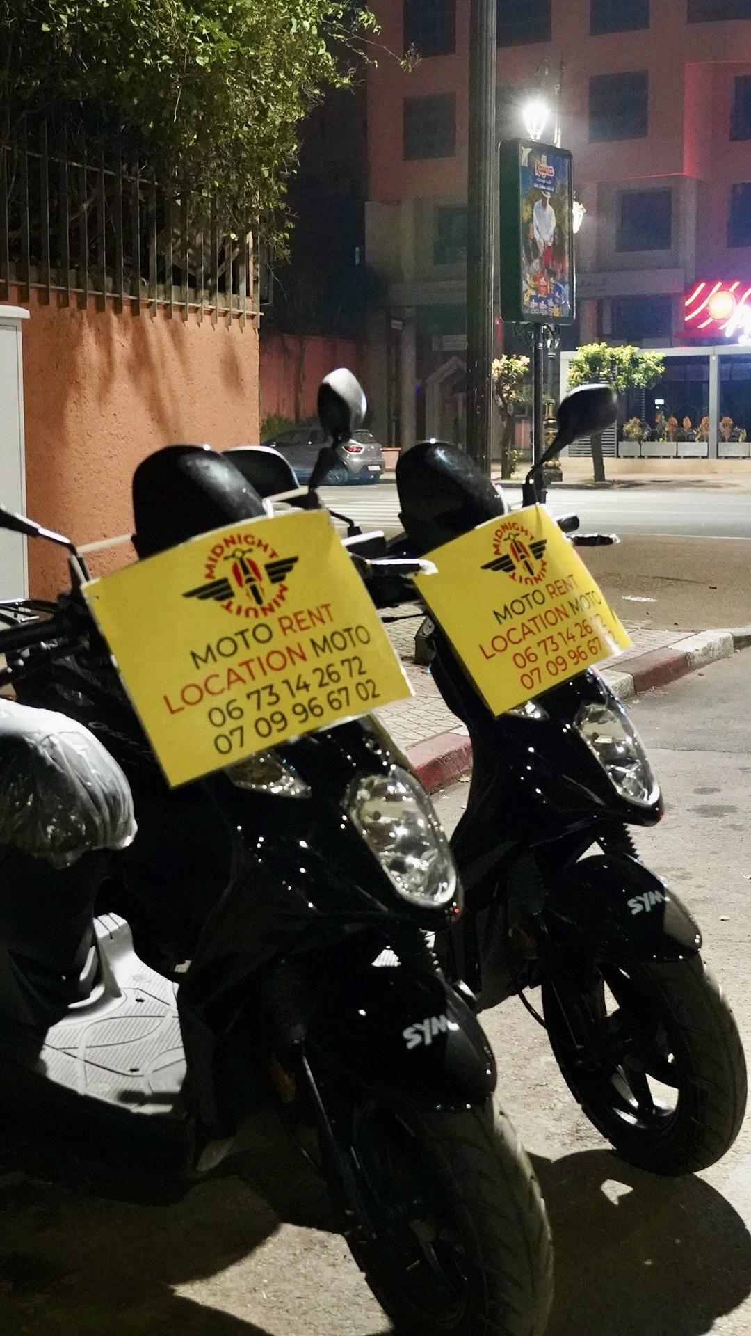 Scooters Minuit Midnight vue face avec sacoches Moto Rent et numéros de contact, sous les néons de Marrakech
